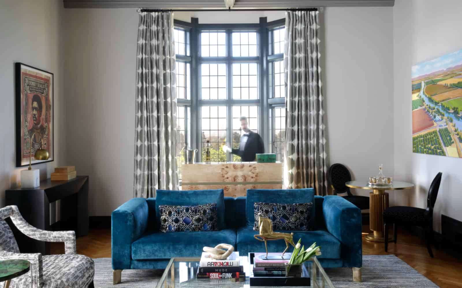 A historical home, the Morse House, in Oakland features a teal blue velvet couch and floor-to-ceiling curtains in front of a dramatic window. Behind the couch is a bar scene, with a tuxedo-clad bartender blurred in motion.