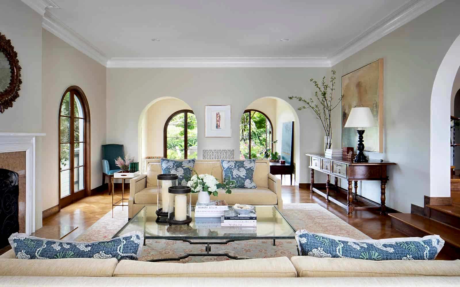 A living room features an arched doorway on the left, two arched doorways along the back wall, and an arched doorway entrance. In the room are two pale yellow couches facing each other in front of a fireplace, with a glass coffee table between them holding candlesticks, books, and a petite flower arrangement.