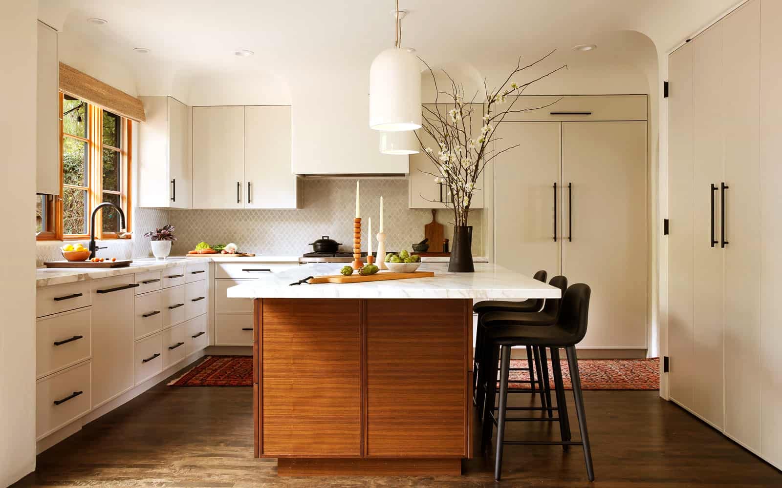 A Mediterranean kitchen features light cream cabinetry, a white marble slab across the counters and the center island, a sink on the left underneath windows framed in warm wood, and a cooking range along the back wall. The island's base is warm toned wood, and sitting atop is a bowl of artichokes and a tall deep brown vase filled with cherry blossom branches.