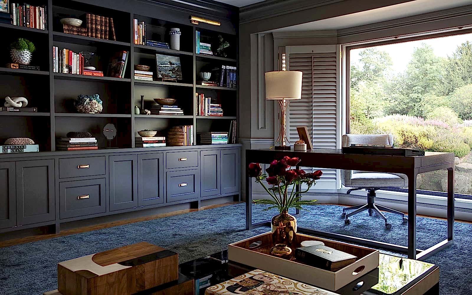 The interior of an office features built-in deep plum bookshelves on the left are adorned with books and trinkets, with an elegant desk in front of a large window on the right and a coffee table with deep red roses in the foreground. 