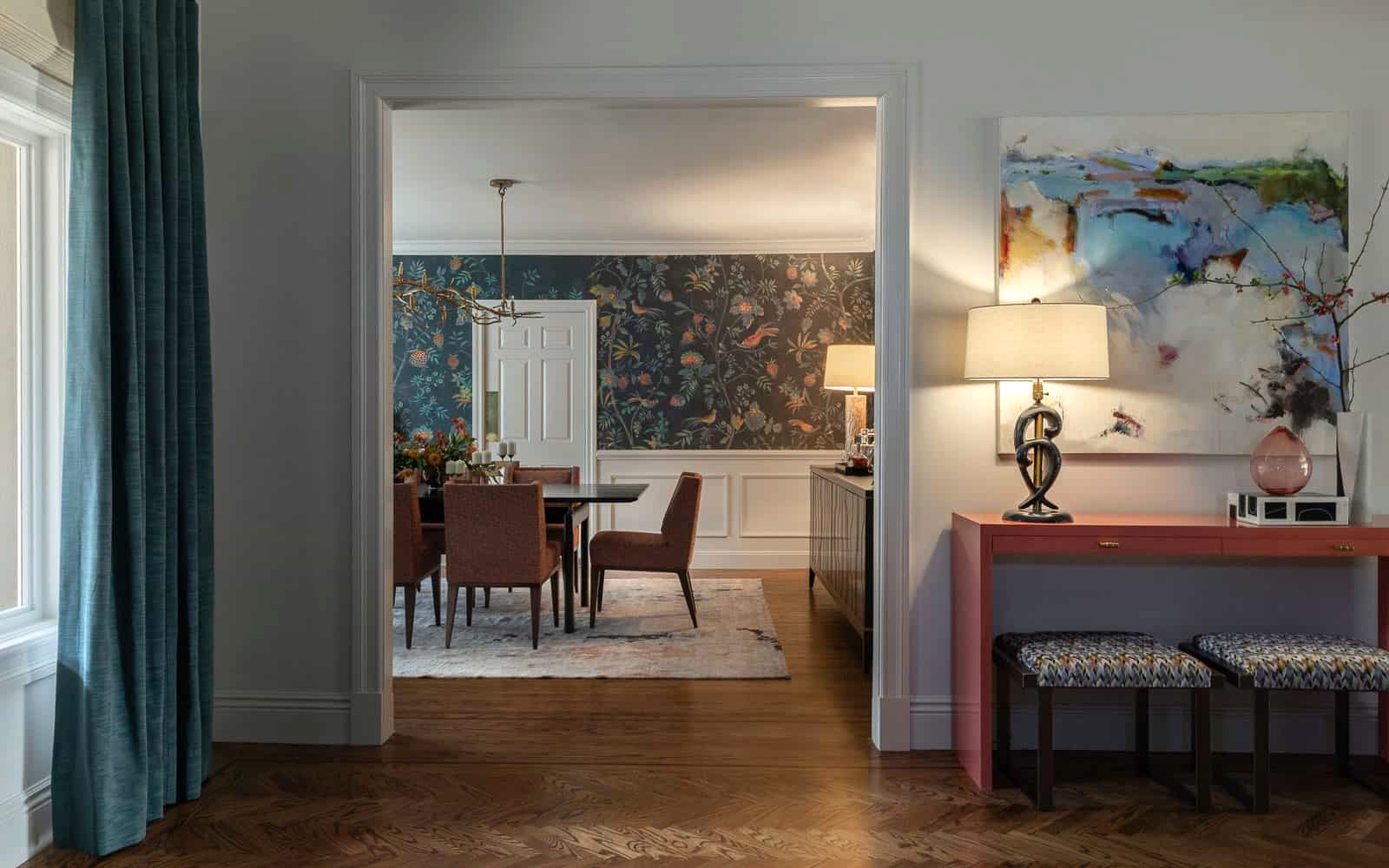 A wide doorway gives a glimpse into an elegant dining room with burnt orange patterned chairs and walls featuring dark blue wallpaper with colorful birds and flowers. Outside of the doorway on the right is a glossy pink hallway table with a green vintage lamp, and abstract art above.