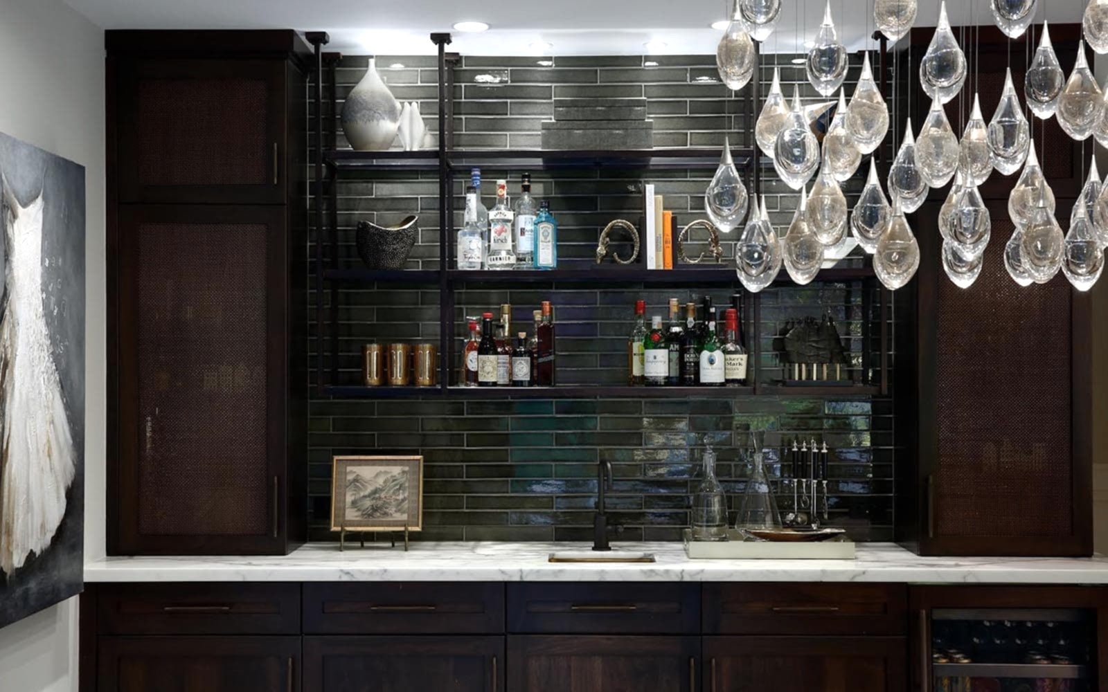 An elegant bar features horizontal rectangle glossy deep green tiles, dark brown cabinets, and a painting of a white dress to the left of it. There is a sink in white marble slab at the wet bar, and drops of clear glass of the room's chandelier are in the foreground.