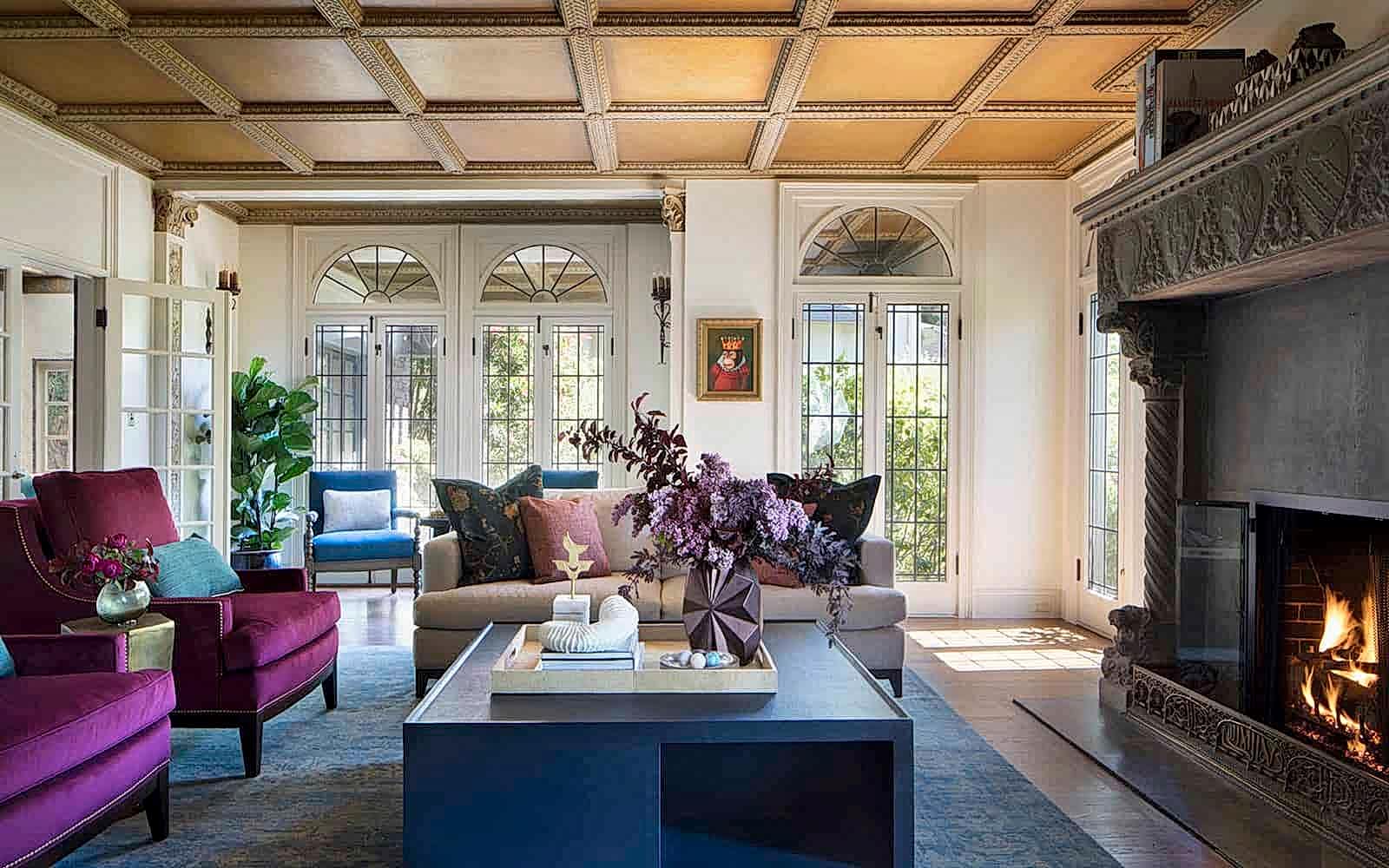 A cozy living room features purple velvet arm chairs in front of a roaring fireplace. The room is sun-filled from the windowed doors, and a coffee table in front of the chairs features a geometric vase with a purple flower arrangement.