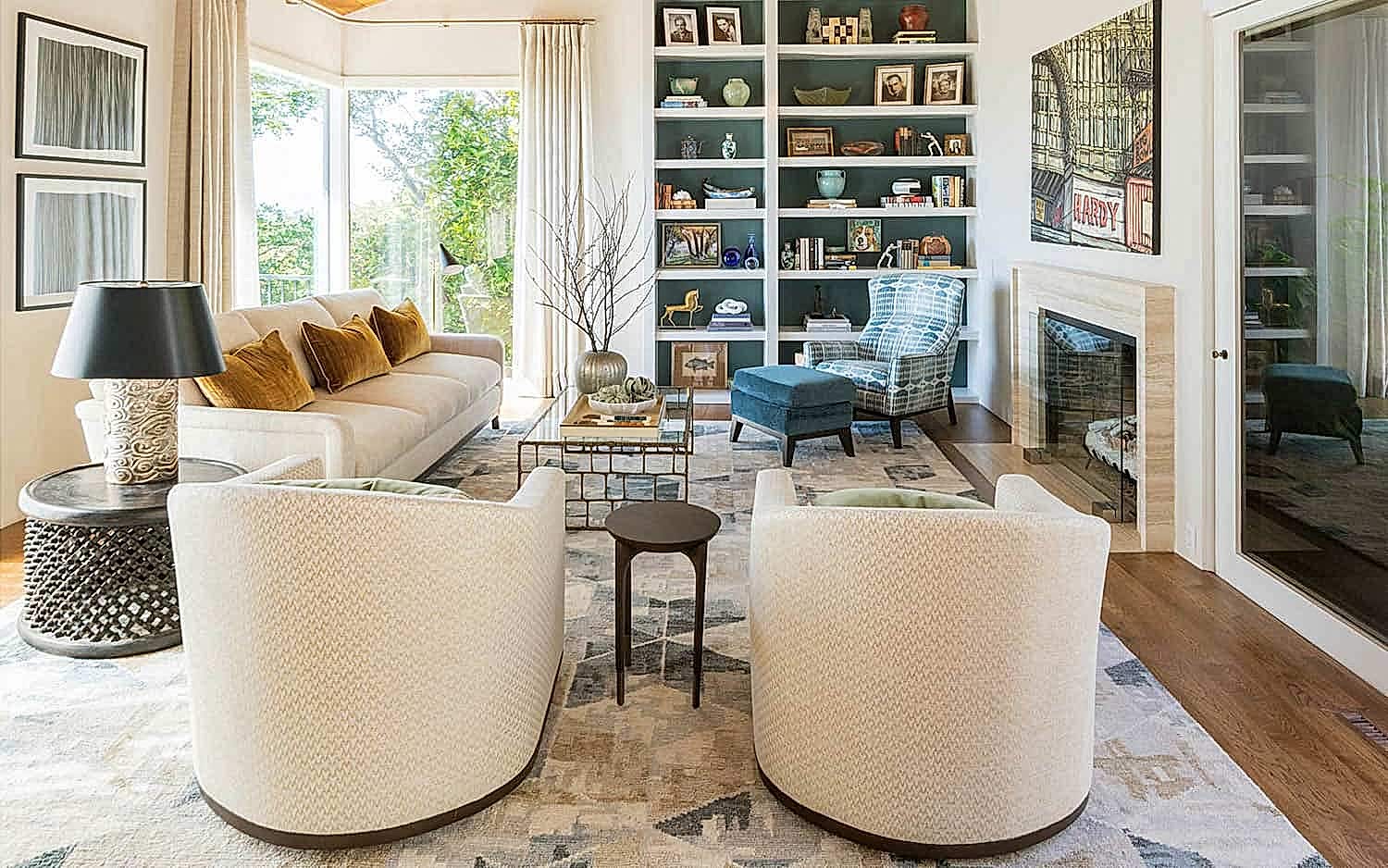 An Oakland Hills remodeled living room is complete with a styled floor-to-ceiling built-in bookcase, a blue corner chair, a creme couch, and a set of 2 matching cream swivel chairs. A corner of the room includes floor-to-ceiling windows that show off the backyard garden.
