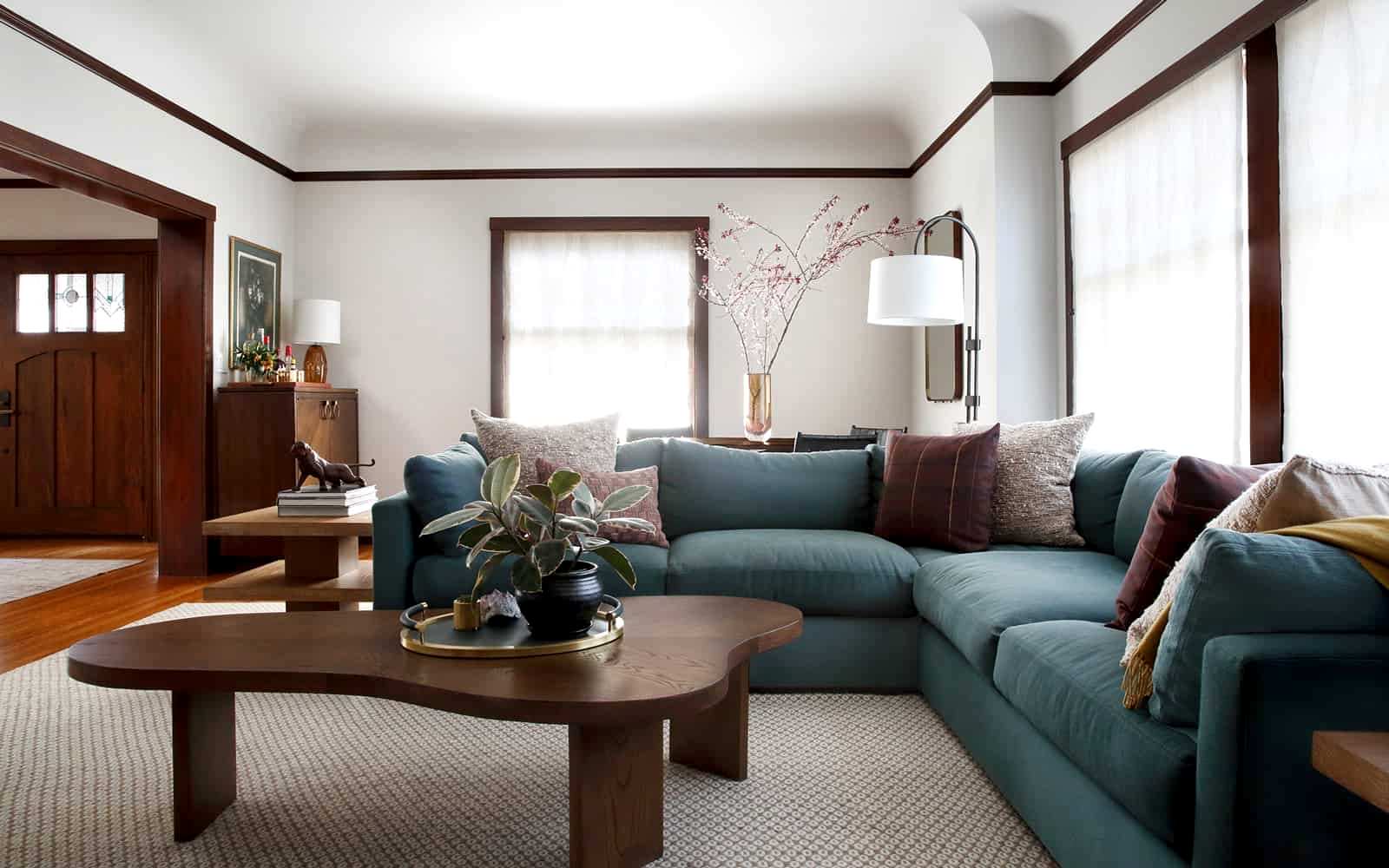 The living room in this Berkeley, California Craftsman style home was designed with sheer window treatments to let in plenty of light. A sectional couch bends around a curved wood coffee table, topped with a round tray and a plant.
